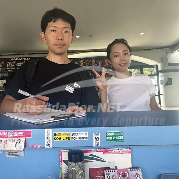 A tourist signing the check-in manifest at a blue Rassada Pier ticket counter, while his companion makes a peace sign, with destination stickers for Koh Lipe and Koh Phi Phi visible.